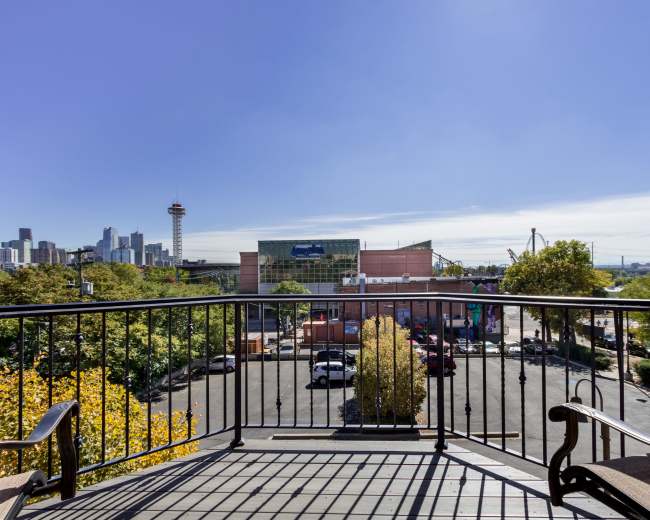A balcony overlooks a cityscape featuring buildings, trees, and parked cars under a clear sky.