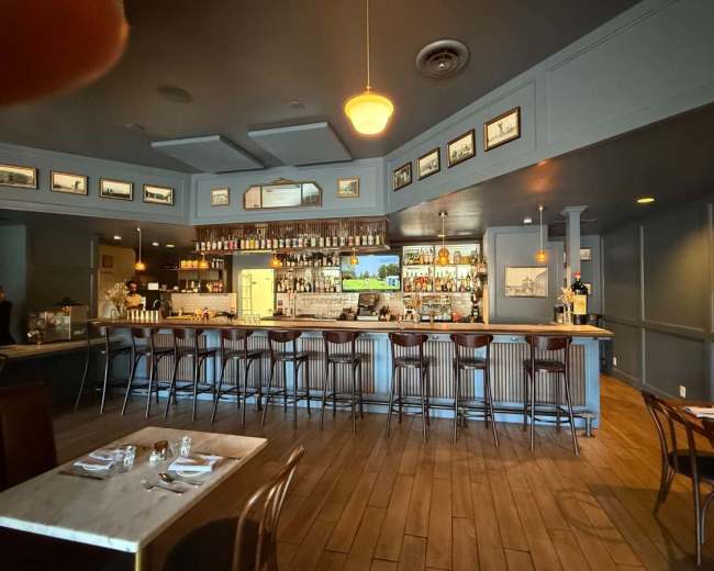 The image shows a dimly lit restaurant bar area with a marble countertop, tall wooden stools, and framed pictures on the walls.