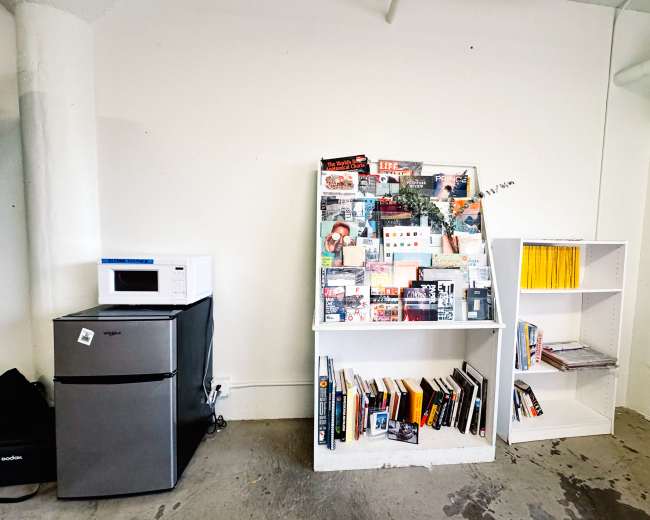 The image shows a room with a microwave and refrigerator on the left and two white bookcases displaying books and magazines on the right.