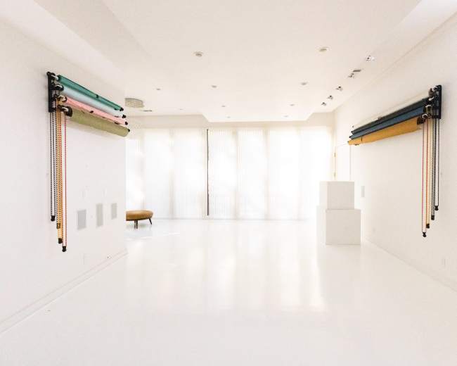 A minimalist room with white walls and flooring features two sets of colored fabric rolls mounted on the walls and large windows with sheer curtains.