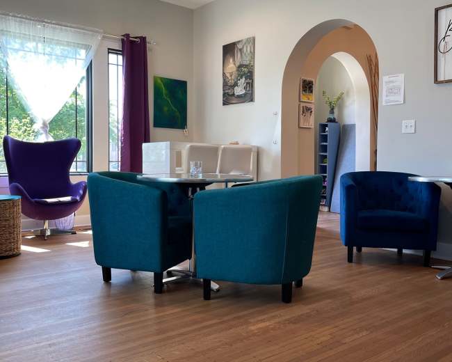 A cozy indoor space featuring two teal armchairs and a glass table, with a purple chair in the background and large windows allowing natural light to enter.