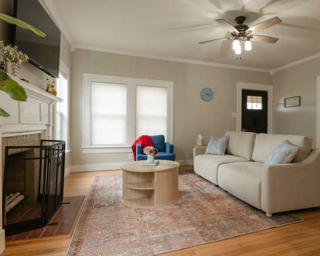 The room features a light-colored sofa, a round wooden coffee table, and a blue armchair, with a fireplace and large windows in the background.