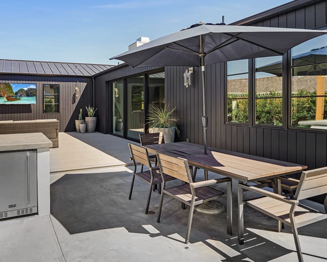 The image shows a modern outdoor patio with a dining table, chairs, and an umbrella, adjacent to a sleek black house and landscaped greenery.