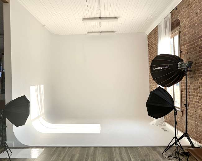 The image shows a photography studio featuring a seamless white backdrop, two softbox lights on tripods, and natural light streaming through a window.
