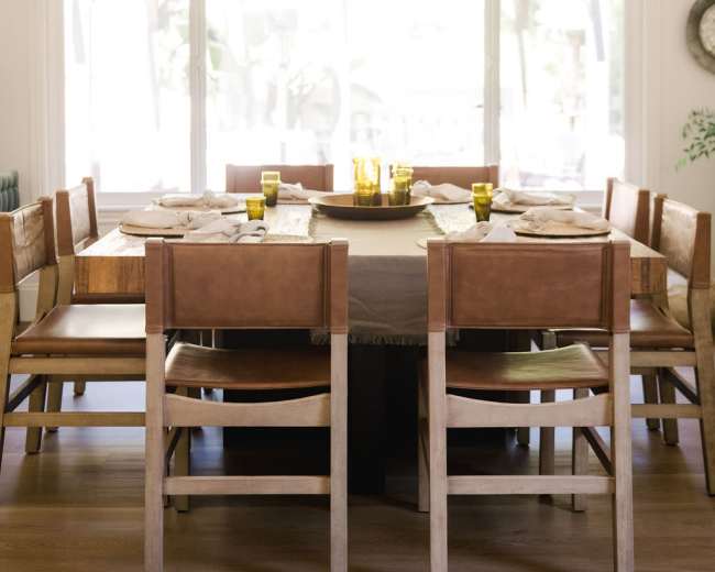 A large wooden dining table surrounded by leather and wooden chairs is set for a meal, with green glasses and plates arranged around it in a well-lit room.