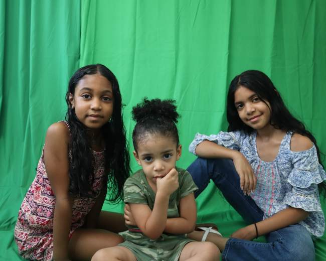 Three young girls pose in front of a green backdrop, with one sitting in the center and the others on either side.