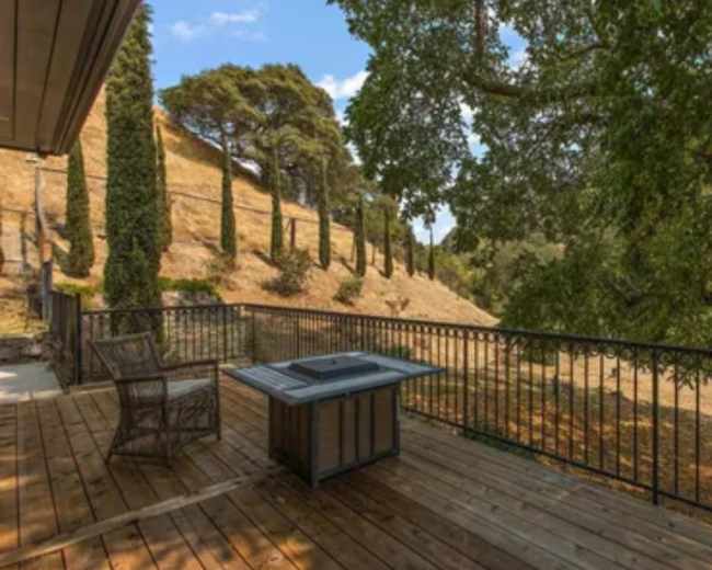 A wooden deck features a table and a chair, overlooking a sloped landscape lined with trees and a clear blue sky.