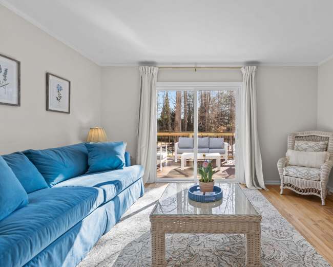 The image shows a cozy living room with a blue sofa, a glass coffee table, and a sliding door leading to a deck area.