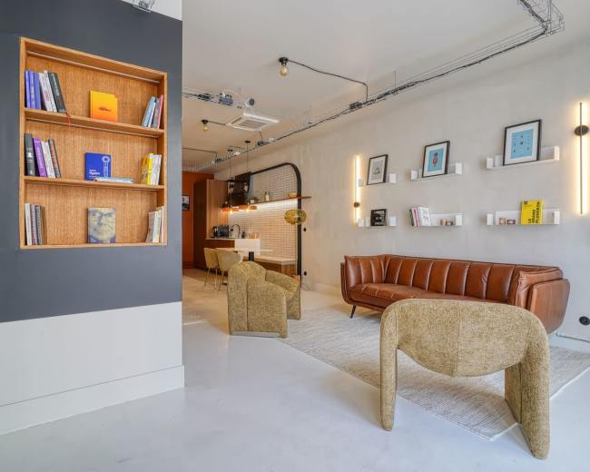 The image shows a modern interior space featuring a brown leather couch, two beige chairs, a wooden bookshelf filled with books, and minimalistic decor on gray walls.