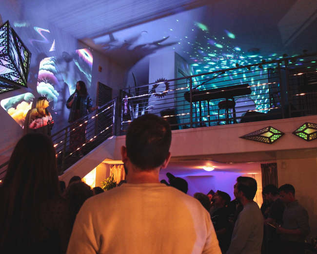 A group of people gathers in a well-lit room featuring projected visuals on the walls and ceiling, with one person speaking from an upper level.