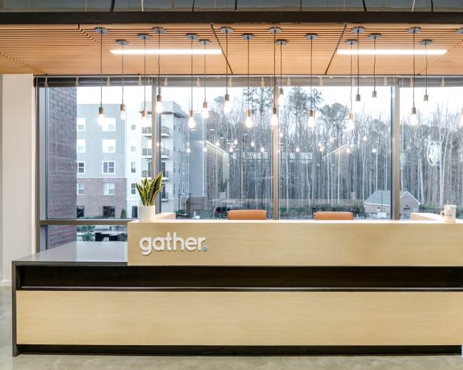 A modern reception desk with the word "gather" is situated beneath pendant lighting, facing large windows that overlook a wooded area.