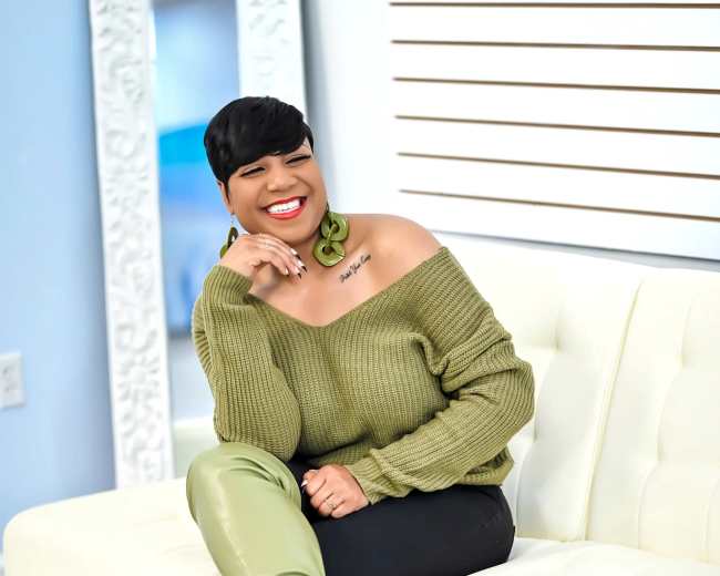A woman wearing a green off-the-shoulder sweater and long green boots smiles while sitting on a white couch.