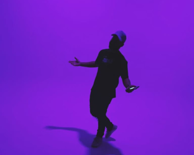 A person stands under a purple background while holding a phone and gesturing with their hands.