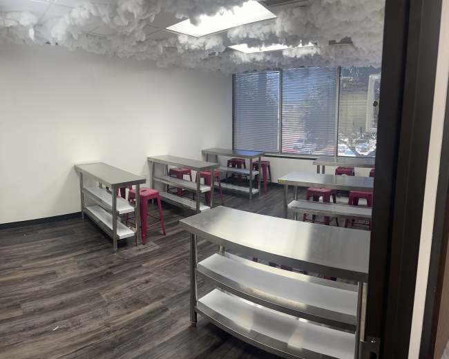The image shows a classroom with several metal tables placed in rows and pink stools, under a ceiling adorned with cloud-like decorations.