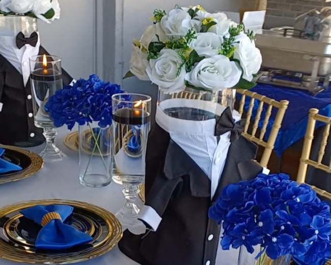 The image shows a dining table set with black, gold, and blue decor, featuring tuxedo-themed centerpieces, blue flowers in glass vases, and plates with blue napkin accents.