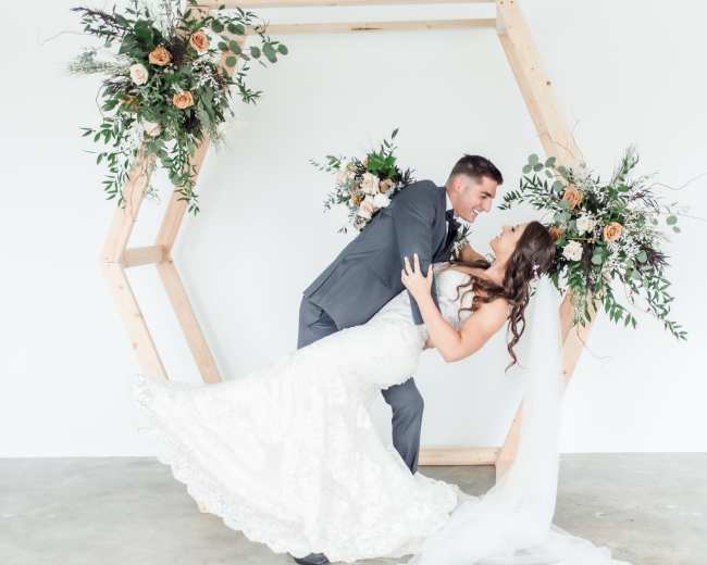 A couple poses in a dip under a wooden hexagonal arch decorated with flowers.