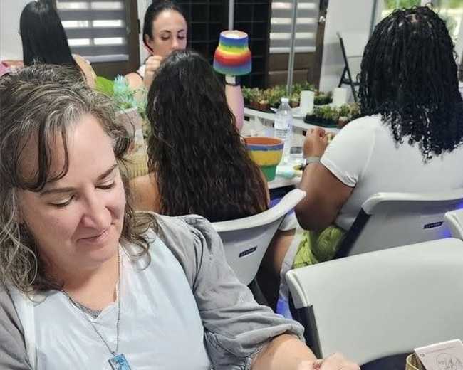 A woman with long hair paints a flower pot at a table surrounded by other participants in a crafting workshop.
