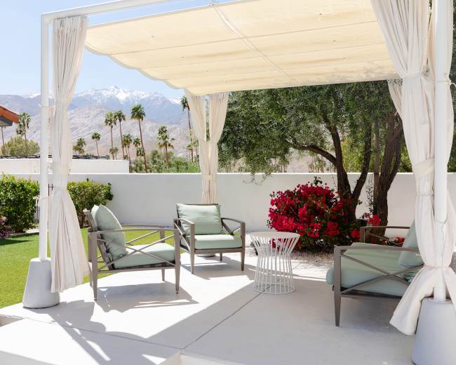 A shaded seating area with four chairs and a small table is surrounded by greenery and colorful flowers.