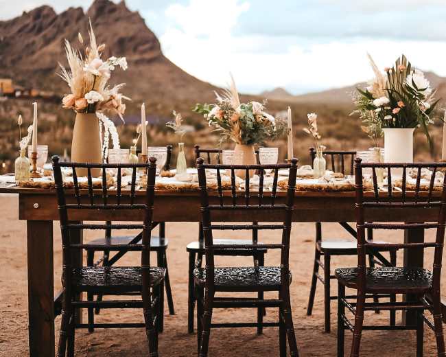 A rustic outdoor dining table is set with floral arrangements, candles, and chairs, surrounded by desert mountains.
