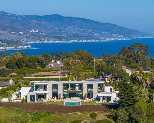 A modern coastal mansion with expansive windows overlooks the ocean and surrounding hills.