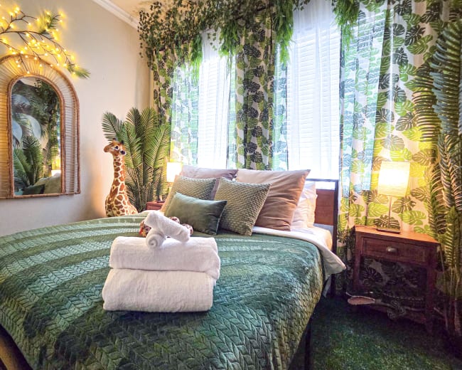 The image shows a cozy bedroom decorated with green bedding, a giraffe statue, and plants, illuminated by warm fairy lights and natural light from the windows.