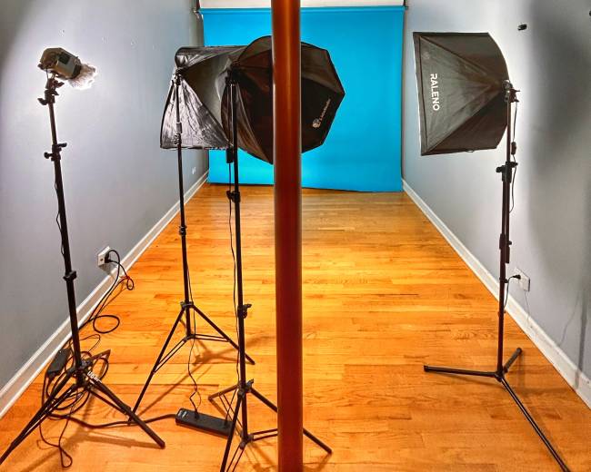 The image shows a photography studio setup featuring a central support pole, two light stands with softboxes, and a blue backdrop against a wooden floor.