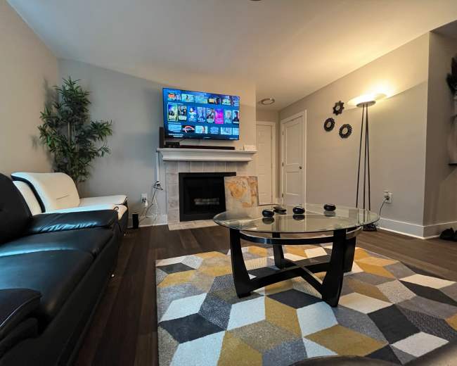 The image shows a cozy living room with a black leather sofa, a glass coffee table on a patterned rug, and a wall-mounted TV above a fireplace.