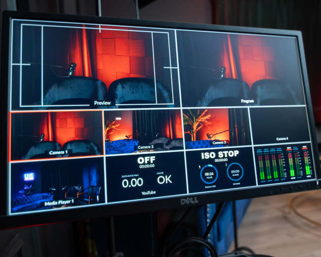 The image shows a computer monitor displaying multiple camera feeds and a media player interface in a studio setting.