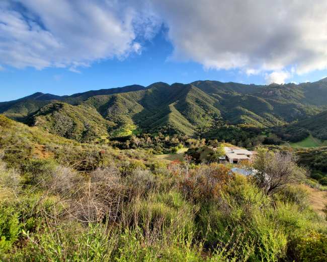 A green, hilly landscape with a distant house nestled among the mountains under a partly cloudy sky.
