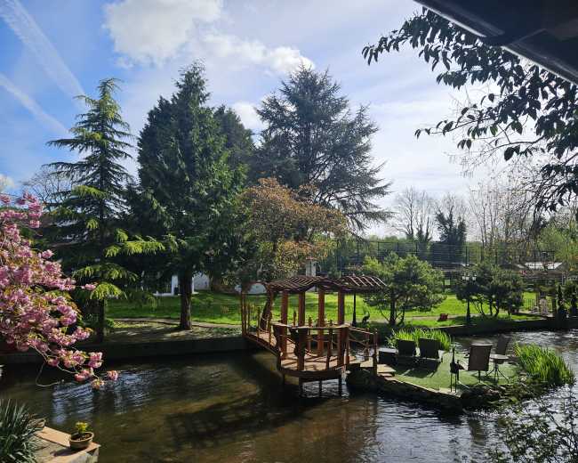 A wooden gazebo is situated on the bank of a tranquil stream, surrounded by lush greenery and flowering trees under a partly cloudy sky.
