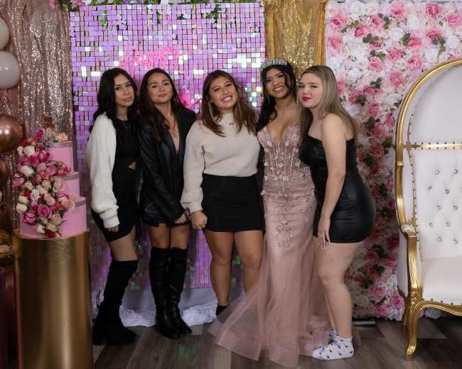 A group of five women poses together in a decorated party area featuring balloons, a floral backdrop, and a cake display.