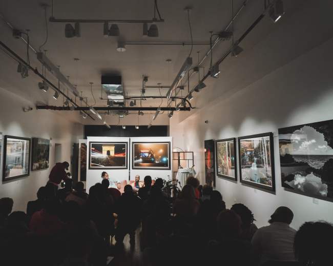 A group of people sit in an art gallery, facing a screen displaying a video, surrounded by large framed photographs on the walls.