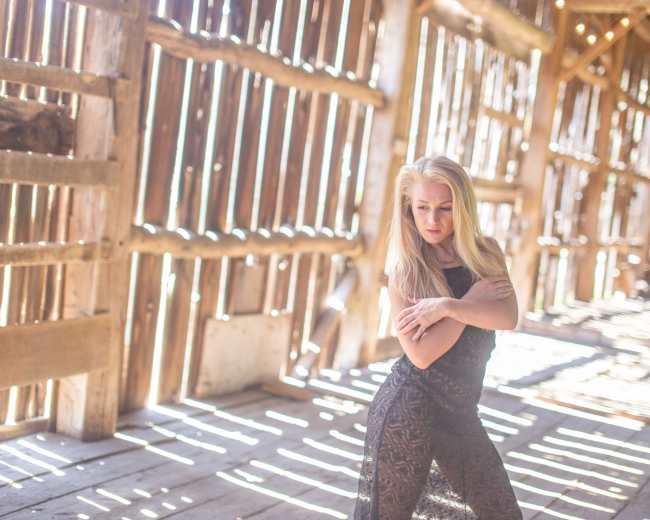 A woman poses in a rustic barn with sunlight streaming through the wooden slats, casting shadows on the floor.
