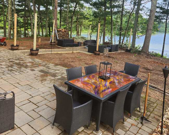 A patio area with a wooden table and chairs is set among trees near a lake, featuring stone pathways and various planters.