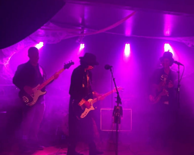 A band performs on stage with a misty atmosphere and purple lighting, while pumpkins are displayed in front of them.