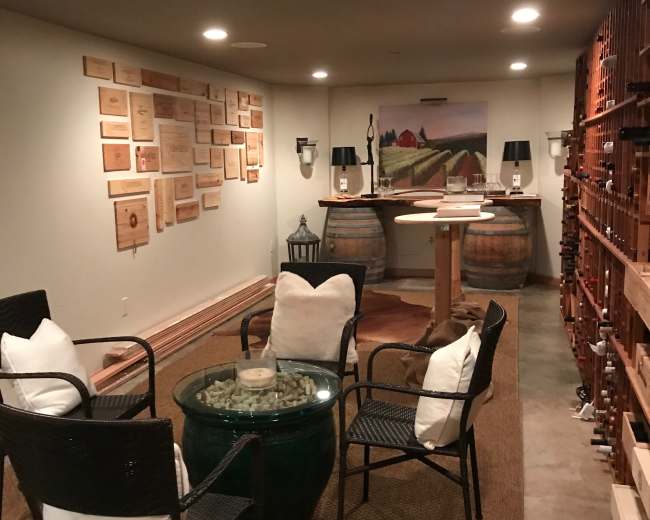 A cozy wine cellar featuring a seating area with four chairs surrounding a glass table, wooden barrels, and wine racks lined along the walls.