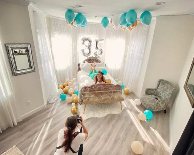 A photographer captures a woman sitting on a bed decorated with balloons and a "35" balloon display in a well-lit room.
