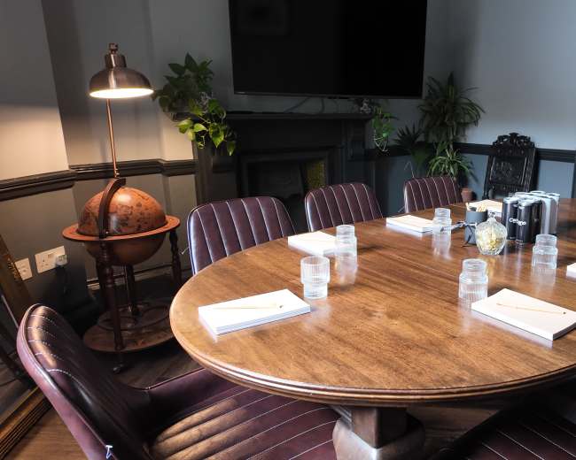 A large wooden table is set with notebooks and glasses of water, surrounded by leather chairs in a dimly lit conference room.