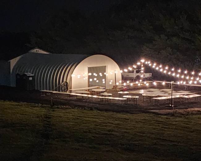 An illuminated outdoor gathering area with string lights and tables is set up near a large, curved building at night.