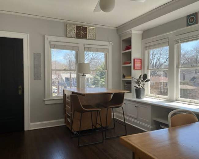 A dining area features a wooden table and chairs beside large windows that overlook a neighborhood.