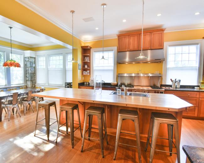 The image shows a spacious kitchen with wooden cabinets, a large island with bar stools, and a dining area featuring a set of tables and chairs.