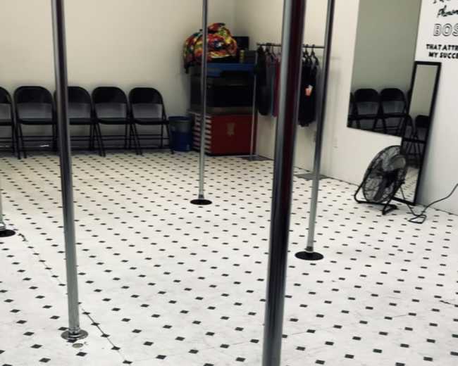 The image shows an empty room with multiple shiny pole dancing poles on a tiled floor, surrounded by folding chairs and a mirror along one wall.