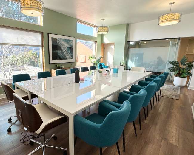 A large, modern conference room with a white table surrounded by teal and white chairs, featuring large windows and plants.
