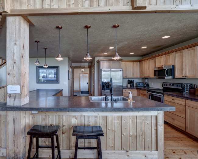 The image shows a modern kitchen with wooden cabinetry, stainless steel appliances, and a bar counter with two stools.