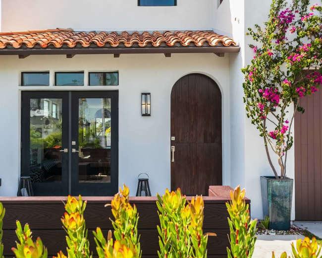 A modern home with a red tile roof, dark wooden doors, and a surrounding garden featuring green plants and a flowering bush.