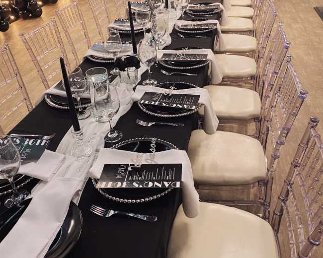 The image shows a long dining table set with black and white tablecloths, elegant glassware, black candles, and floral centerpieces in a decorated room.