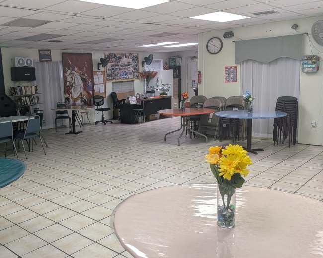 The image shows an interior space with multiple tables and chairs, featuring flower arrangements on the tables, and a service area or office visible in the background.
