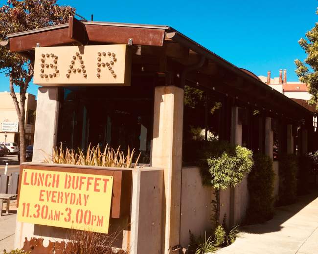 A building with a large sign reading "BAR" and a poster advertising a lunch buffet is located on a clear day with a blue sky.