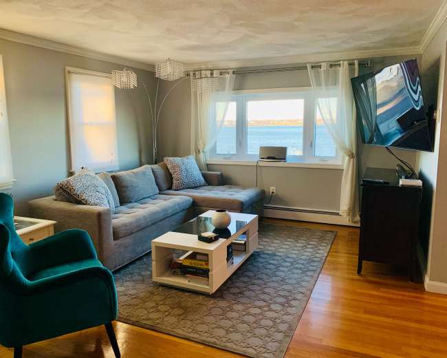 The living room features a gray sofa and an armchair, with a coffee table and a television, overlooking a water view through large windows.