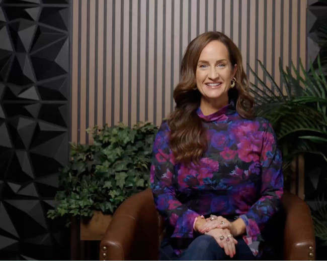 A woman with long hair sits on a brown chair in front of a striped wall and green plants.
